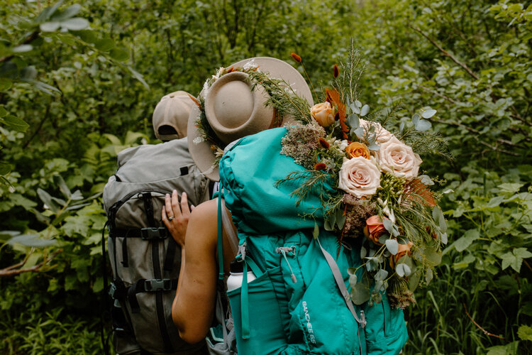 Eloping couple with backpacks hike through green trails in Glacier National Park